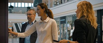 Es sind zwei Frauen und ein Mann zu sehen. Sie betrachten gemeinsam etwas und sprechen darüber.