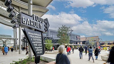 Auf dem Bild ist ein Totempfahl mit der Aufschrift &quot;Food Garden&quot; zu sehen, dahinter stehen Gebäude.