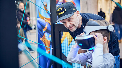 Ein kleines Kind mit VR Brille und der Vater mit einer Cap mit Batman Logo.
