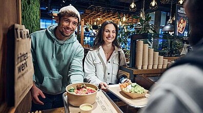 Eine Frau und ein Mann holen sich ihr Essen, einen Salat und eine Portion Pommes bei frittenwerk ab.