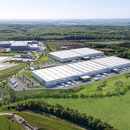 Vogelperspektive auf eine weite grüne Wiese und mittendrin ein großes Logistikgebäude.