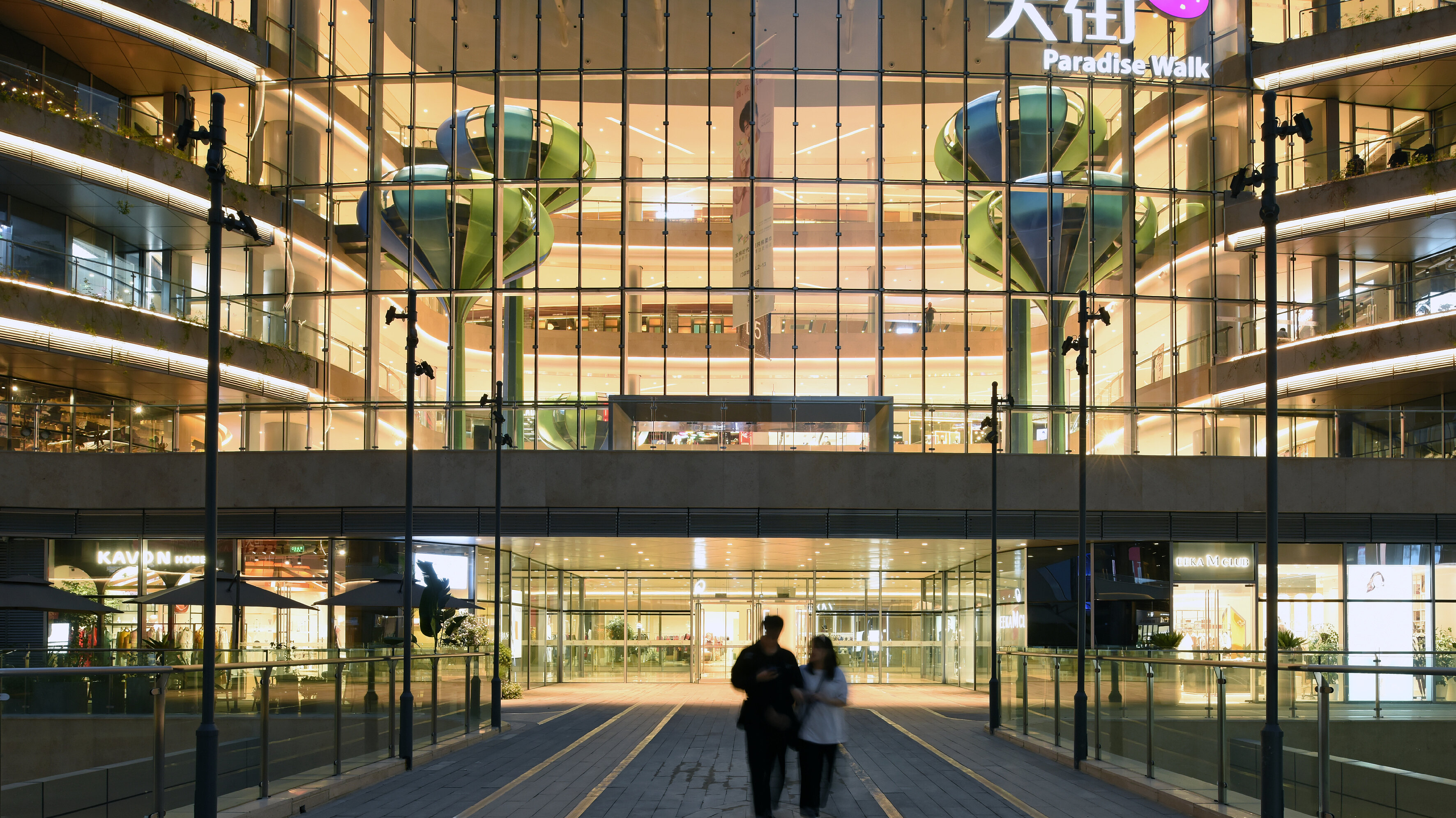 View of the brightly lit entrance area of a shopping centre with a Chinese-language logo on the façade, under which the words &#039;Paradise Walk&#039; can be read.