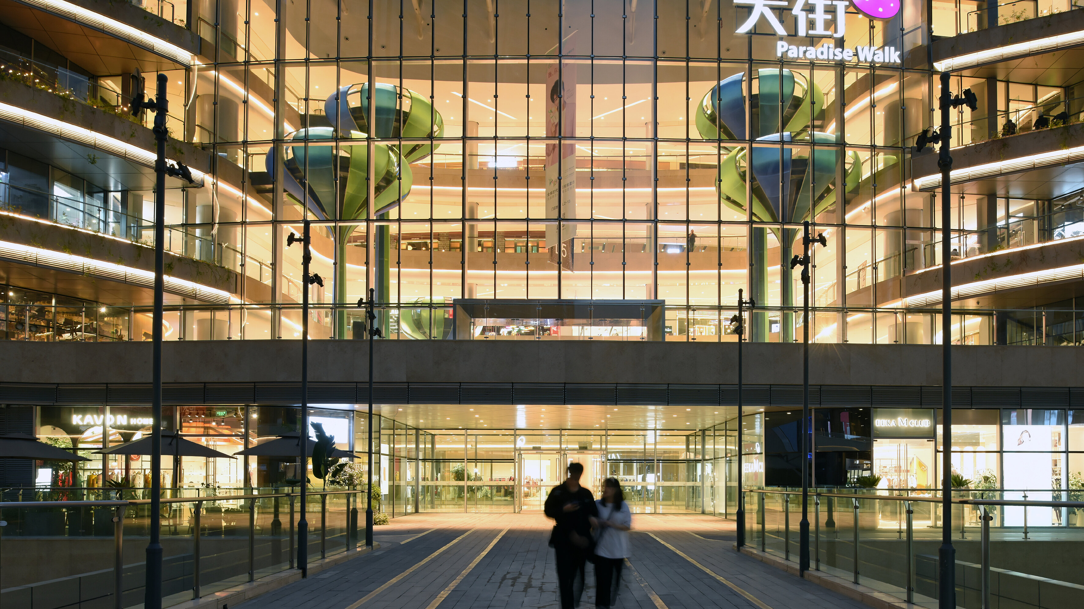 Blick auf den hell erleuchteten Eingangsbereich eines Shopping-Centers mit einem chinesischsprachigen Logo an der Fassade, unter dem der Schriftzug ‚Paradise Walk‘ zu lesen ist.