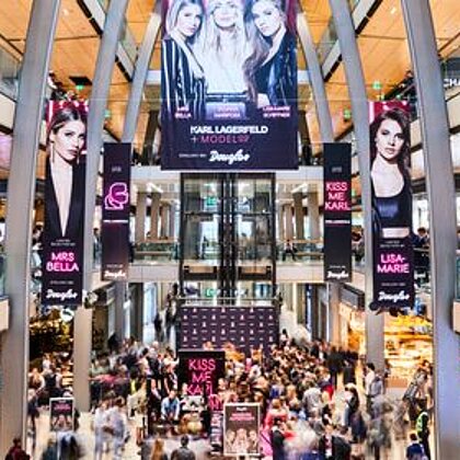 Inmitten eines Shopping-Centers hängen vier große Plakate von der Decke, die für ein Event werben