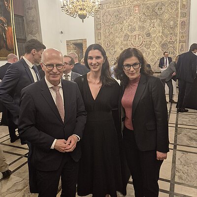 Drei Personen in formeller dunkler Kleidung lächeln freundlich in die Kamera.