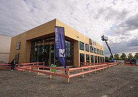 Auf dem Bild ist ein großes Gebäude zu sehen, mit Baukran und Baustellenzaun.