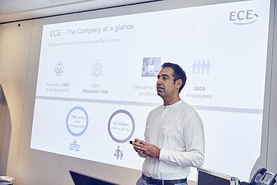 A person in a white shirt stands in front of a screen while a presentation slide is shown in the background