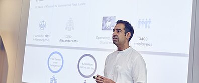 Eine Person steht vor einer Leinwand, im Hintergrund wird eine Präsentationsfolie gezeigt