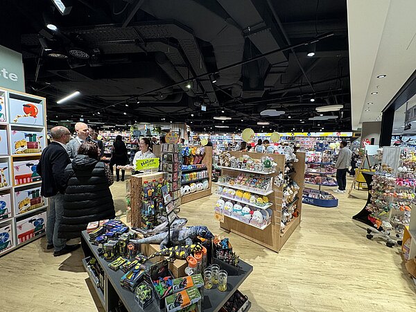 Ein gut beleuchteter Verkaufsbereich mit mehreren Regalen voller Spielwaren und Geschenkartikel, in dem einige Menschen zwischen den Auslagen stöbern.