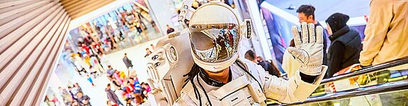 Eine Person in einem Astronautenanzug befindet sich in einem rege besuchten Shopping-Center und winkt in die Kamera.
