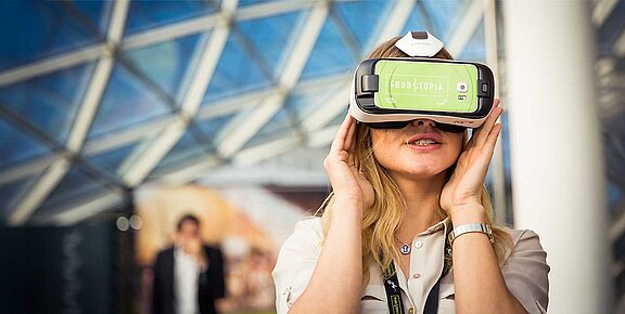 A woman looks around through VR goggles, an impressive glass dome can be seen in the background.