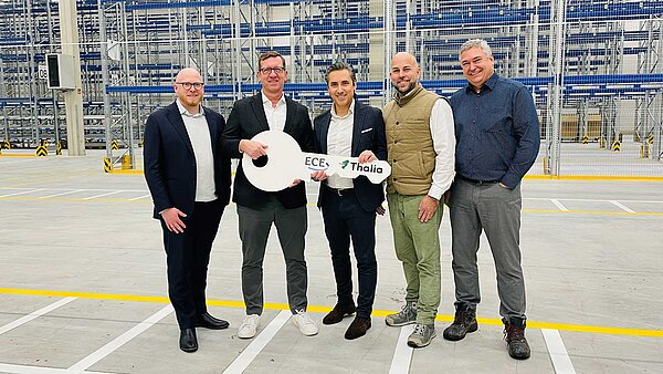 Auf dem Bild sind fünf Männer in einer Lagerhalle zu sehen, zwei von ihnen halten einen großen weißen Schlüssel in der Hand, auf dem das Logo von ECE und Thalia abgedruckt ist.