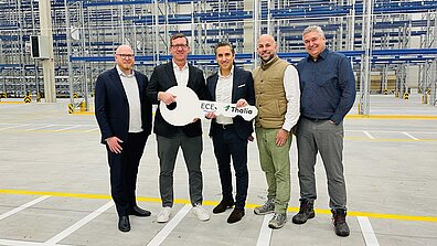Auf dem Bild sind fünf Männer in einer Lagerhalle zu sehen, zwei von ihnen halten einen großen weißen Schlüssel in der Hand, auf dem das Logo von ECE und Thalia abgedruckt ist.