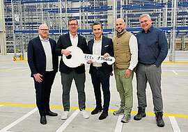 Auf dem Bild sind fünf Männer in einer Lagerhalle zu sehen, zwei von ihnen halten einen großen weißen Schlüssel in der Hand, auf dem das Logo von ECE und Thalia abgedruckt ist.