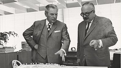 Schwarz-weiß Fotografie mit Werner Otto und einer weiteren Person, die vor einem Bauplan stehen