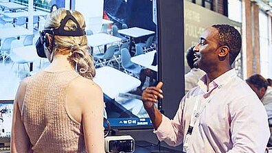 Zwei Personen stehen vor einem großen Bildschirm. Eine der Personen trägt eine VR-Brille.