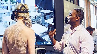 Two people are standing in front of a large screen. One of them is wearing VR glasses.