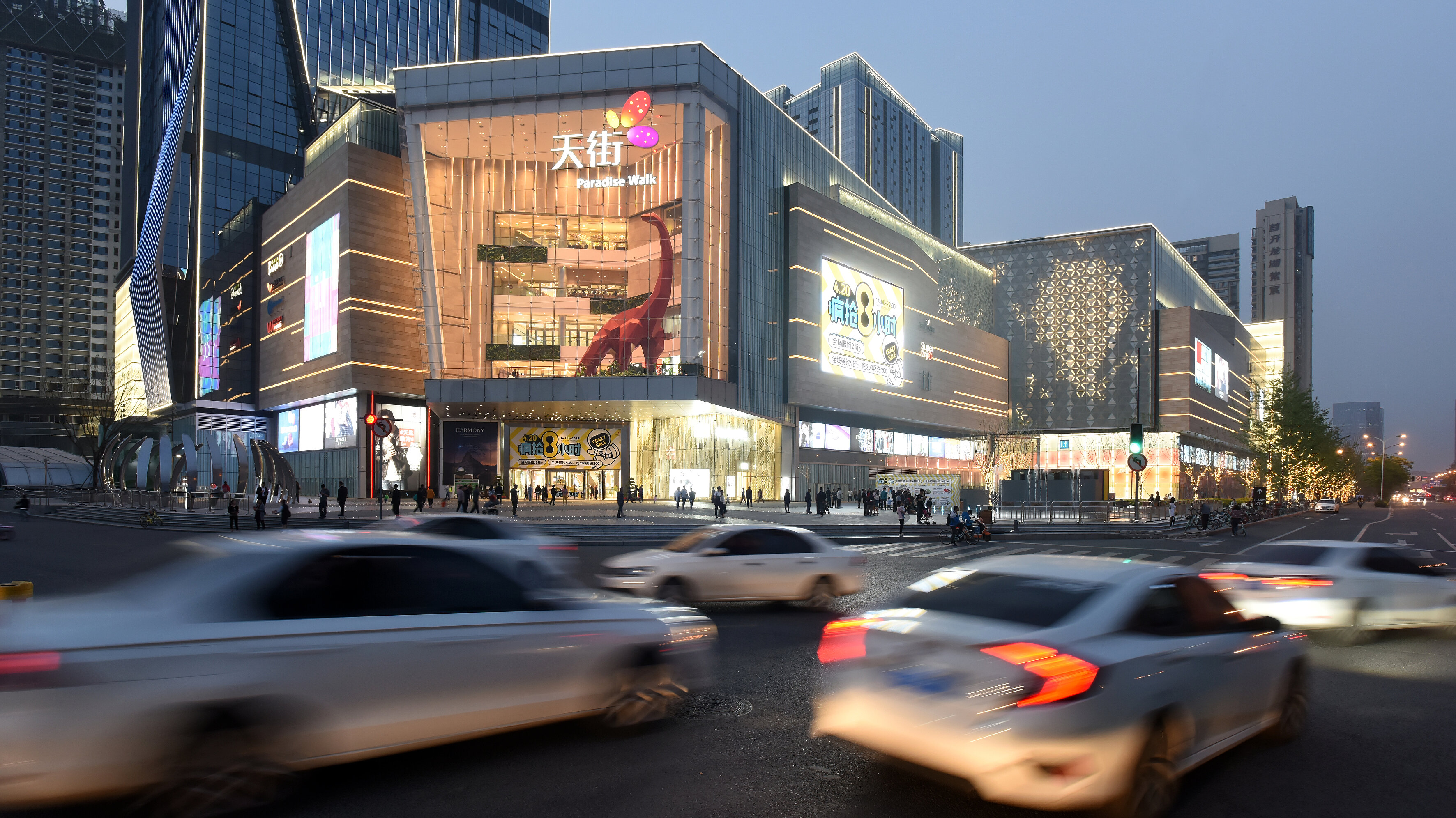 Abendliche Straßenszene vor einem hell erleuchteten Shopping-Center mit großflächigen LED-Fassaden inmitten urbaner Hochhauskulisse.