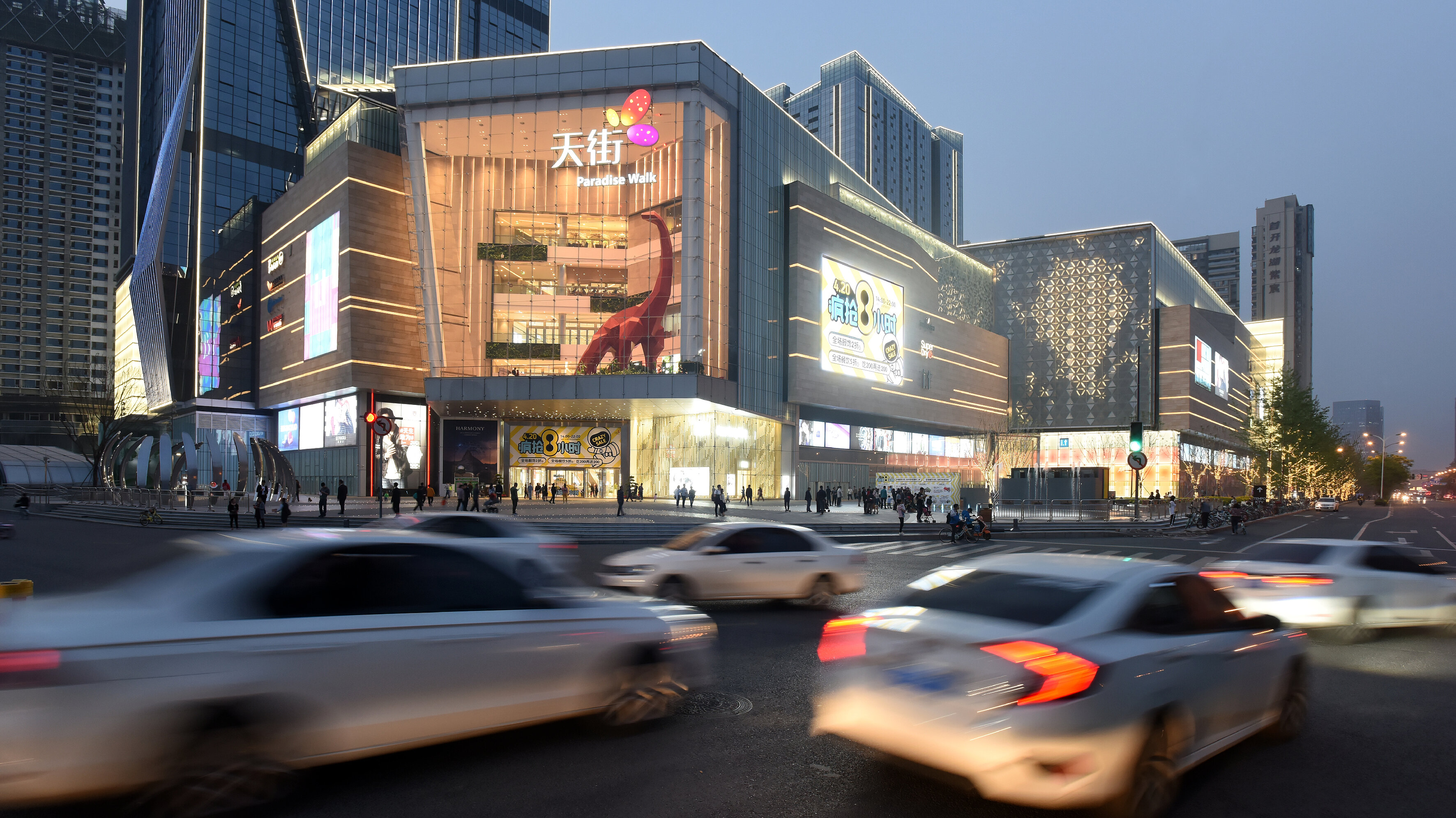 Evening street scene in front of a brightly lit shopping centre with large areas of LED façades amidst an urban backdrop of high-rise buildings.