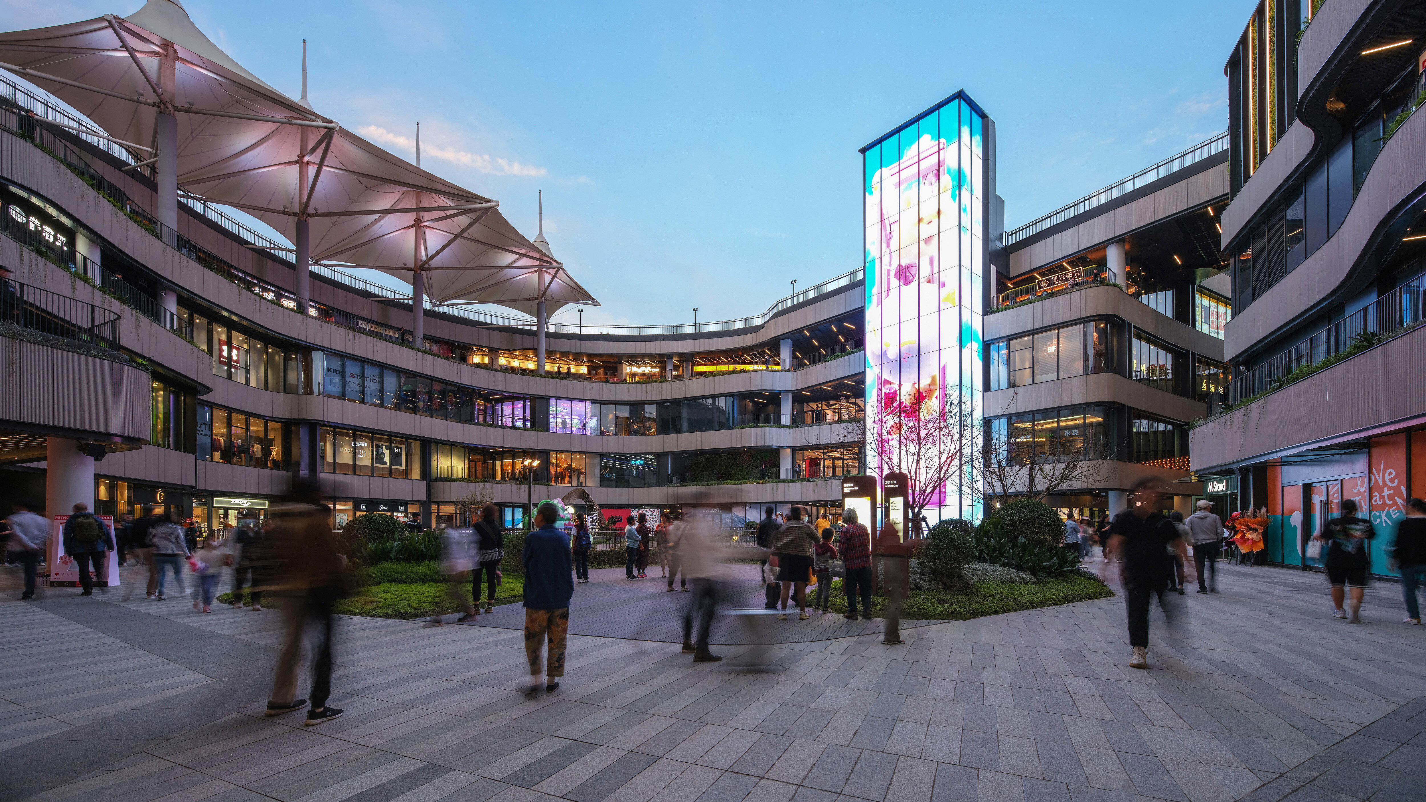 Szene aus dem Außenbereich eines gut besuchten Shopping-Centers, geprägt von geschäftigem Treiben und einer markant beleuchteten Säule.