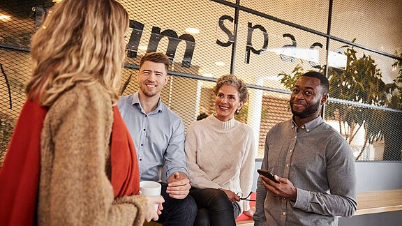 Ein Team aus unterschiedlichen Personen befindet sich in einer Lounge und wirken fröhlich.