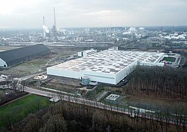 Auf dem Bild ist das Logistikcenter als große weiße Halle zu sehen, dahinter Umrisse eines Industriegebiets.