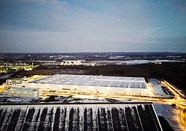 Auf dem Bild ist das beleuchtete Logistikcenter als große Halle zu sehen, dahinter Umrisse eines Industriegebiets.
