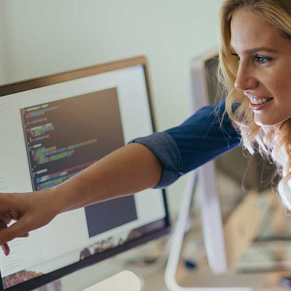 Ein Mitarbeiter sitzt vor Bildschirmen und eine Kollegin steht neben ihm und zeigt auf einen der Bildschirme.