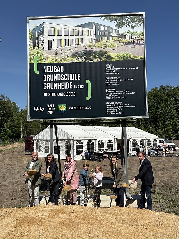 Junge und ältere Teilnehmer setzen gemeinsam zum symbolischen Spatenstich an. Hinter ihnen kündigt ein Plakat den Bau einer neuen Grundschule an.