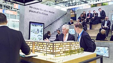 People in respectable black suits are engaged in an exchange. In the foreground, two people are entertained by a large model of a building.