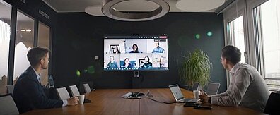 Zwei Männer im Konferenzraum schauen auf einen Bildschirm, der Online-Teilnehmer im Meeting zeigt.