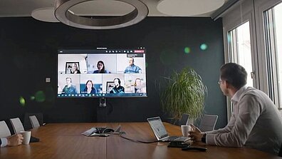 Zwei Männer im Konferenzraum schauen auf einen Bildschirm, der Online-Teilnehmer im Meeting zeigt.