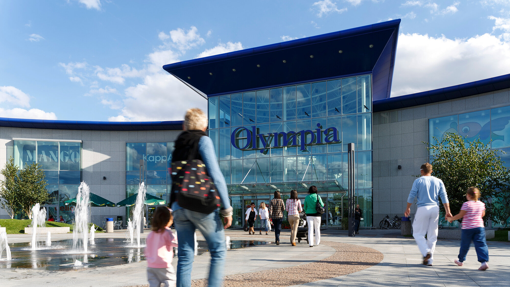 Blick auf den Platz vor dem tschechischen Einkaufszentrum Olympia mit vielen Menschen und Wasserfontänen.
