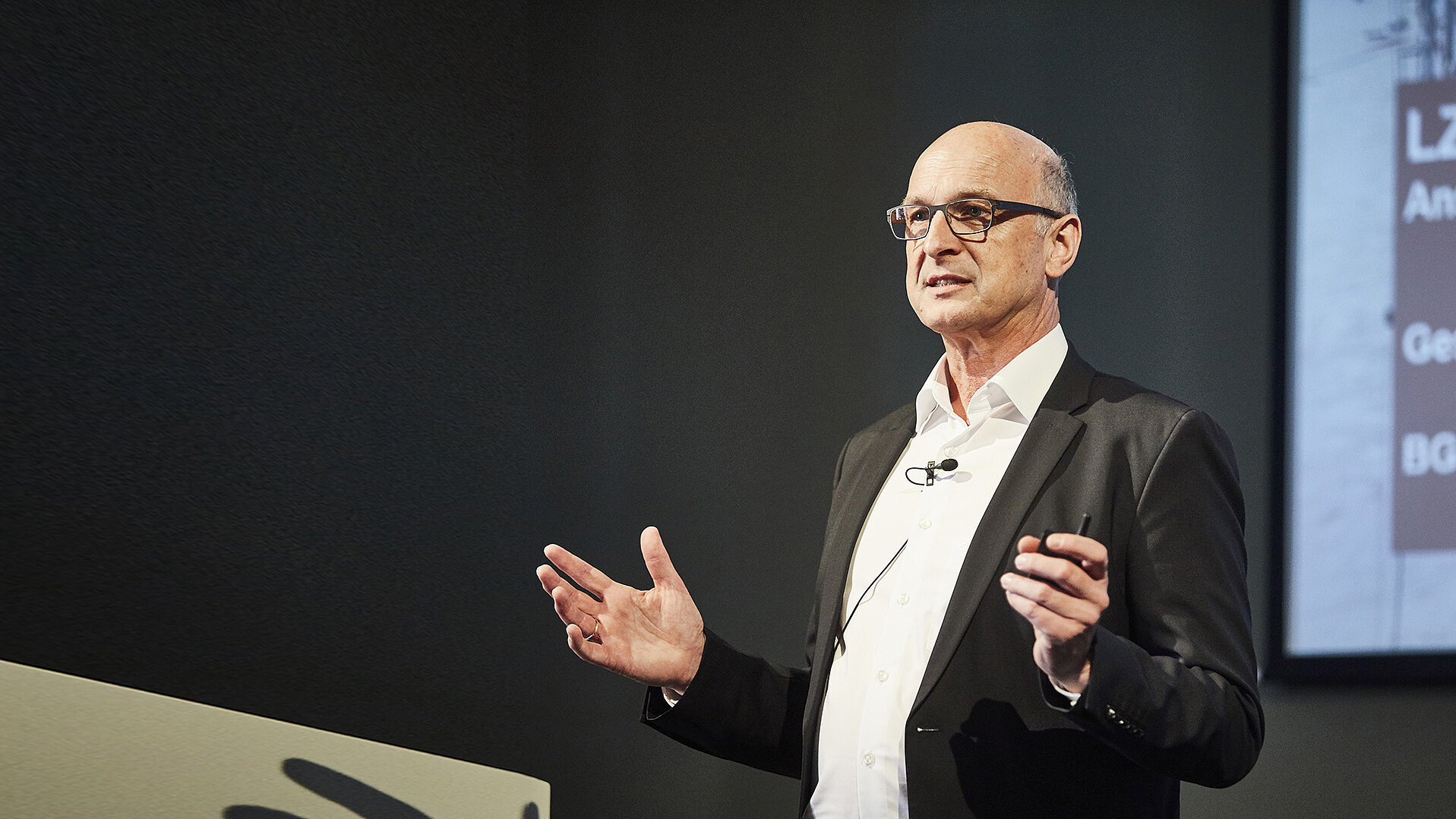 Ein Mann in formeller Kleidung steht mit einem Mikrofon vor einer Leinwand und gestikuliert