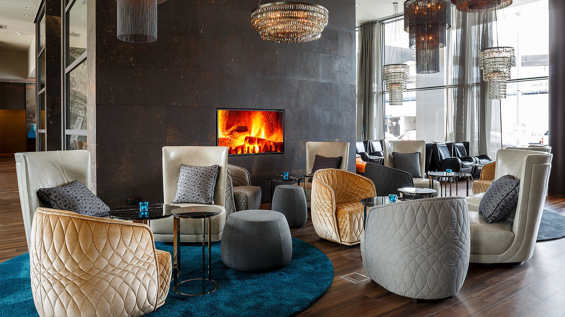 View of a seating area in a hotel with cosy furnishings and an electric fireplace on a dark wall.
