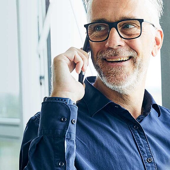 Es ist ein lächelnder, telefonierender Mann in einer hellen Umgebung zu sehen.