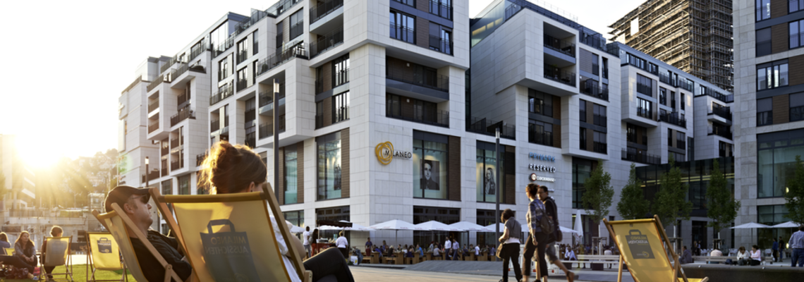 Einige Menschen sitzen bei Sonnenuntergang auf einer Grünfläche und im Hintergrund sieht man moderne Wohngebäude.