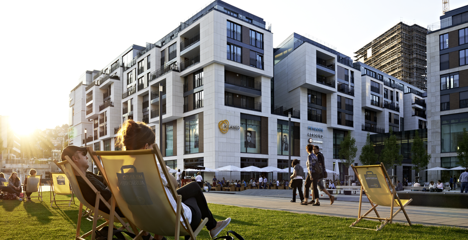 Einige Menschen sitzen bei Sonnenuntergang auf einer Grünfläche und im Hintergrund sieht man moderne Wohngebäude.