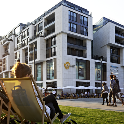 Einige Menschen sitzen bei Sonnenuntergang auf einer Grünfläche und im Hintergrund sieht man moderne Wohngebäude.