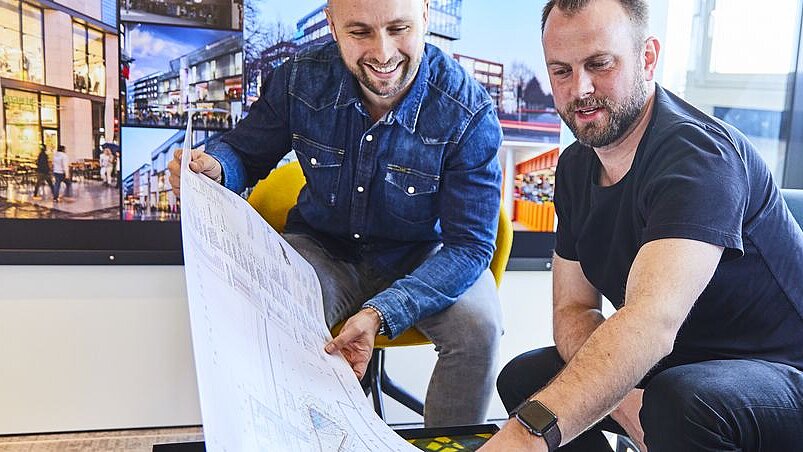 Two people look at an architectural plan on a long piece of white paper