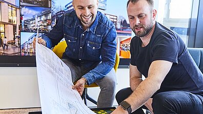 Zwei Personen schauen sich einen Architekturplan auf einem langen weißen Papier an