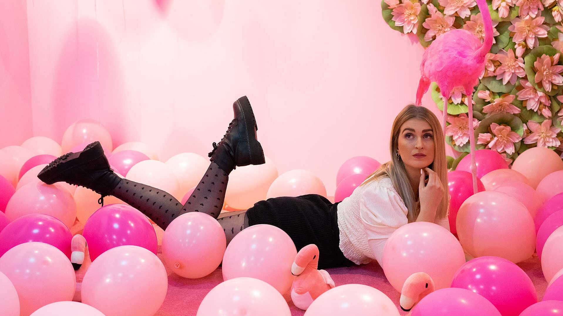 Eine Frau posiert liegend vor einer pinken Wand mit pinken Luftballons.