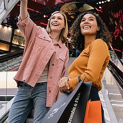 Two women standing on an escalator in a shopping center looking into the distance