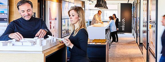 Mehrere Personen befinden sich in einem Arbeitsraum und zwei Personen im Vordergrund arbeiten an einem Architekturentwurf.