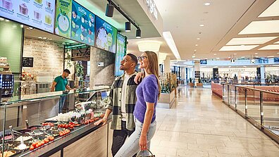 A woman and a man stand in the food court of a center in front of the evergreen offer and look at the screens with the various dishes.