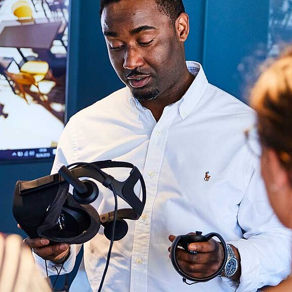 Ein Mann präsentiert zwei weiteren Personen eine VR-Brille