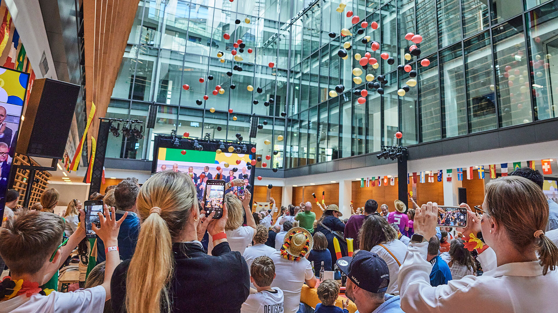 Many people are standing in the ECE atrium watching a soccer match broadcast on large screens and monitors. Many balloons in the colors black, red and gold rise from the ceiling.