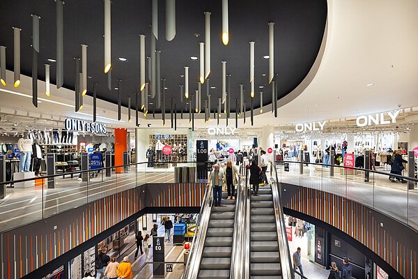 Ein offener Innenbereich eines modernen Kaufhauses mit einer Rolltreppe im Zentrum, umliegenden Verkaufsflächen auf zwei Etagen und vielen beleuchteten Deckenleuchten.