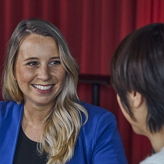 Nahaufnahme einer lächelnden Frau. Rechts sieht man eine weitere Frau von der Seite.