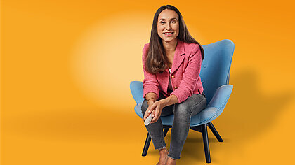 Es ist eine Frau vor einem orangenen Hintergrund zu sehen, die auf einem Stuhl sitzt.